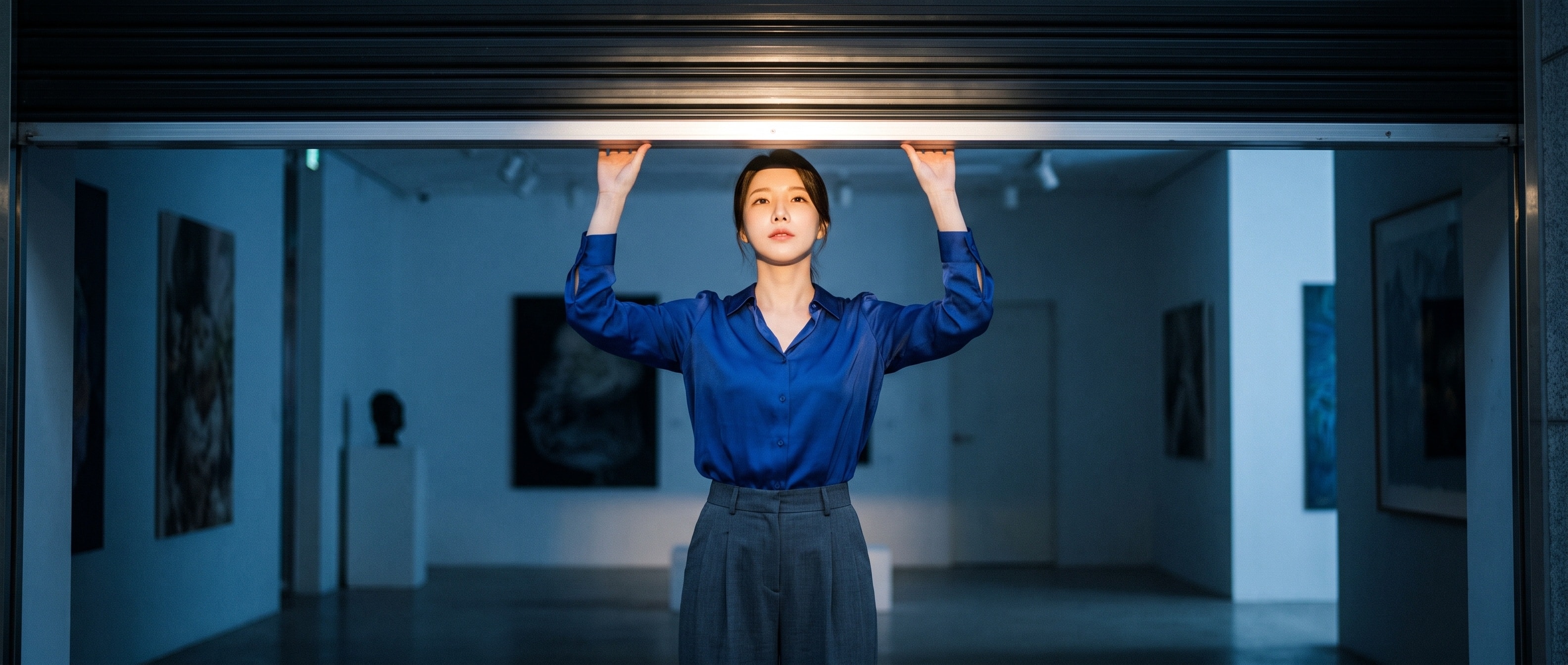 21:9 시네마틱. 어두운 갤러리에서 코발트블루 블라우스에 차콜 슬랙스를 입은 루나가 반쯤 내려온 검은 셔터 아래로 손을 뻗어 그 틈을 다시 조금 들어 올리려 하고 있다. 셔터 뒤로 새어 나오는 빛이 얼굴을 밝힌다. 차분하고 단단한 표정, 얕은 심도, 포토리얼 편집 사진